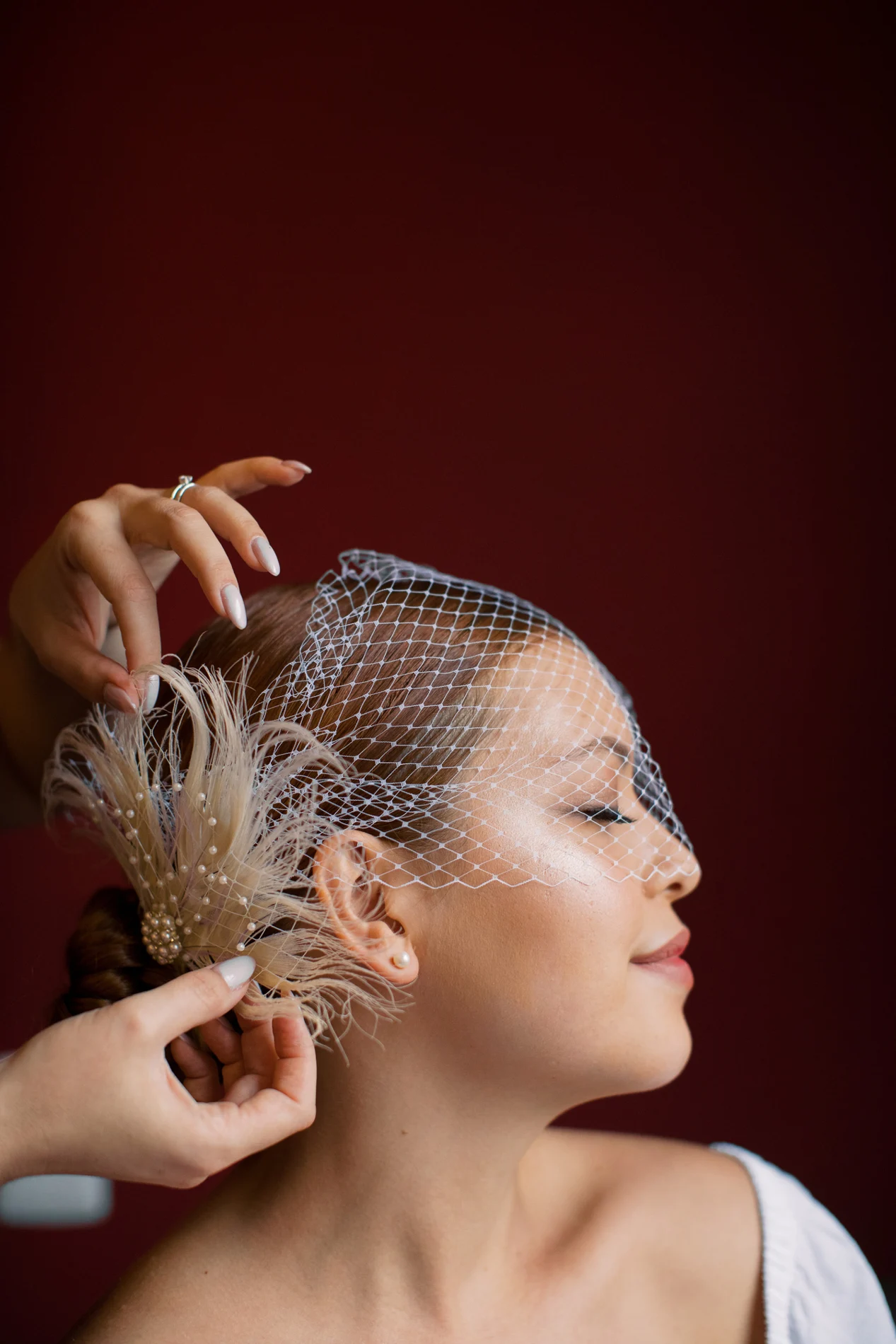 Profile portrait with a birdcage veil being adjusted by hand.